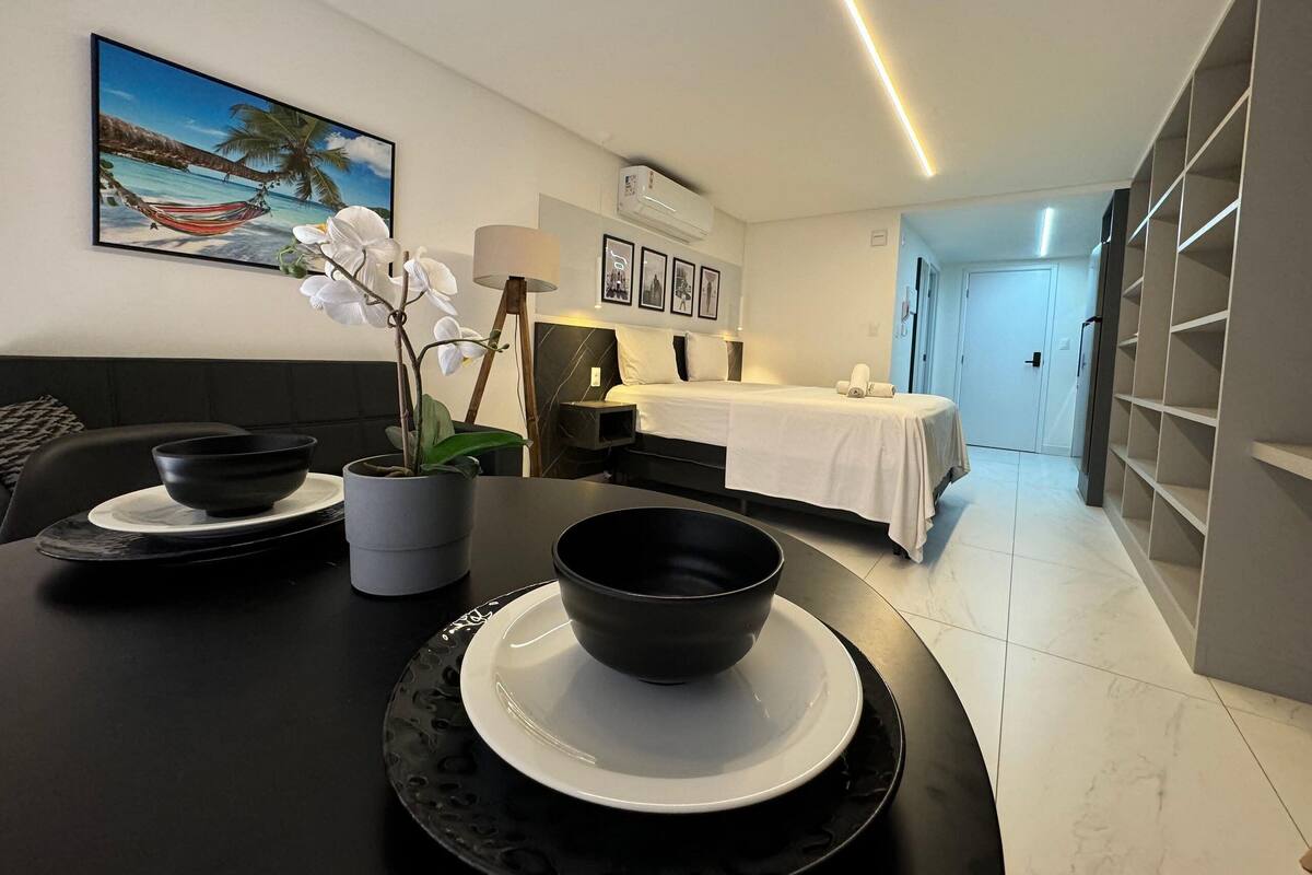 A modern hotel room with a bed, a dining area with a table set for two, a bedside table with a lamp, and a view of the sea through the window.