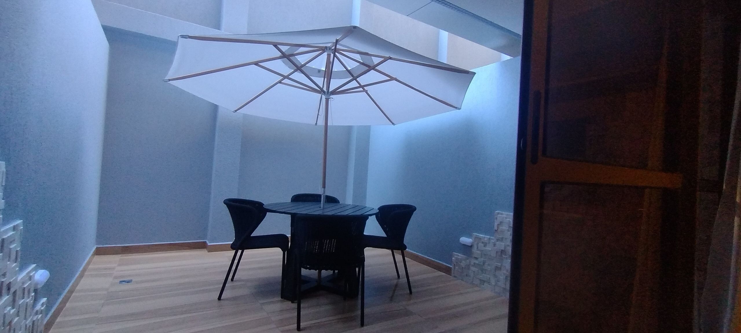A small room with a round table and four chairs, a large white umbrella, and a wooden floor.