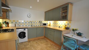 A compact kitchen with a washing machine, microwave, and a small dining area with a chair and table. - 2a Rose St- cosy coastal cottage Fife (Saint Monans)