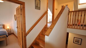 A staircase with wooden steps and a wooden railing. There is a bed with a blue and white checkered blanket and a small bedside table with a lamp. - 2a Rose St- cosy coastal cottage Fife (Saint Monans)