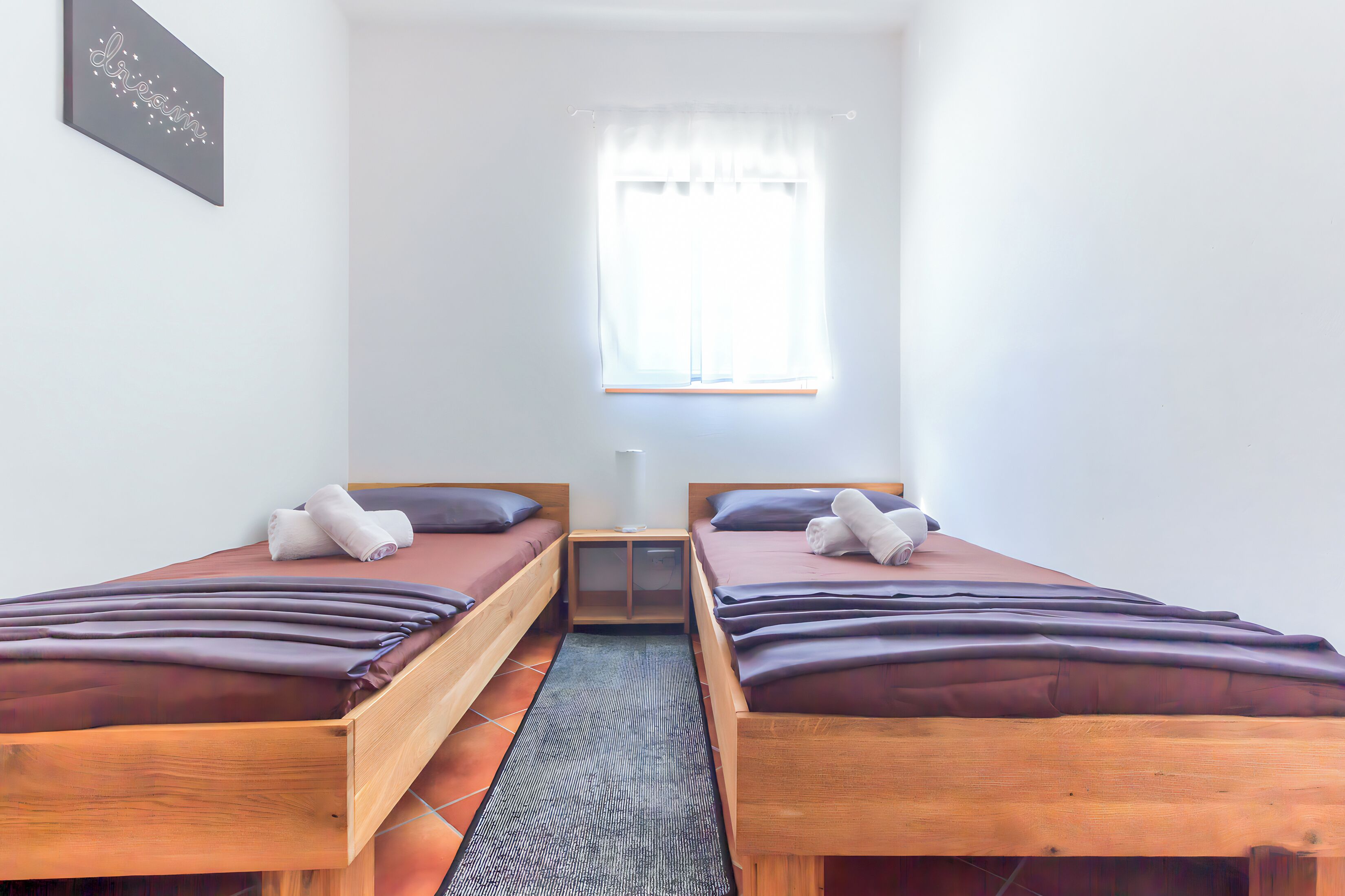 Una habitación pequeña y limpia con dos camas de madera, una ventana con cortinas translúcidas y una mesita entre las camas.