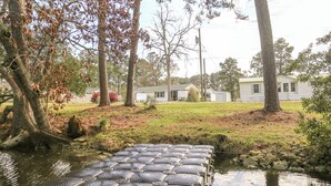 A rustic wooden bridge over a small stream, surrounded by trees and a white building with a green roof. - On the water with kayaks and a launch, 5 mins from beach, Everything you need! (Swansboro)
