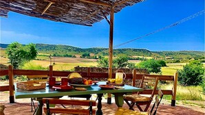 Ein Essbereich im Freien mit einem Tisch, gedeckt für vier Personen, umgeben von Holzstühlen mit gepolsterten Sitzen. Auf dem Tisch befinden sich ein Korb und eine Limonadenkanne.