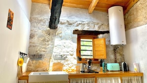 Eine rustikale Küche mit Steinwand, Holzbalken, einem Oberlicht, einer weißen Spüle, einem Holzkasten und einem weißen Warmwasserbereiter.