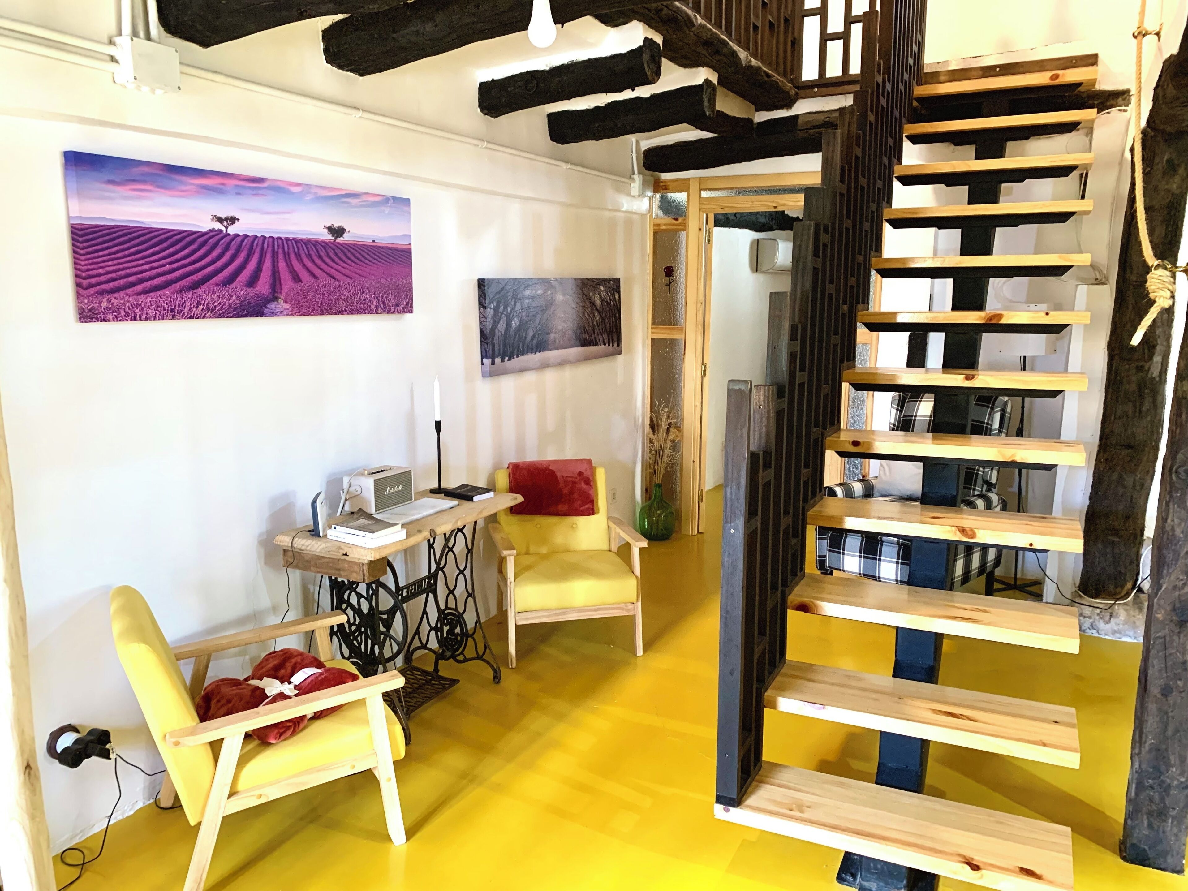 Una habitación con piso amarillo, un sillón amarillo, un escritorio de madera con una lámpara, una estantería y un cuadro de un campo de lavanda.