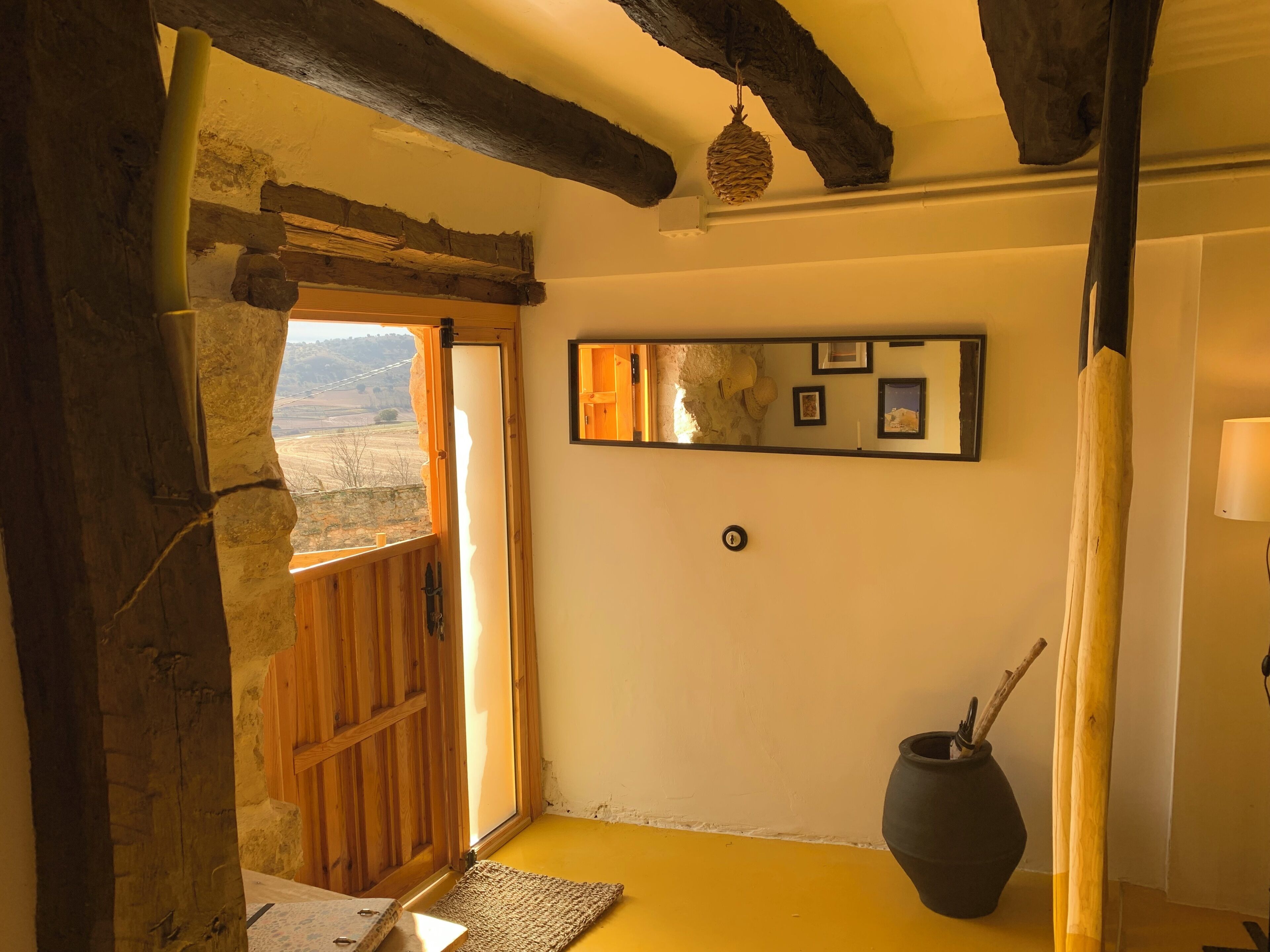 Une pièce rustique avec une porte en bois donnant sur un balcon, un grand vase et un miroir au mur.