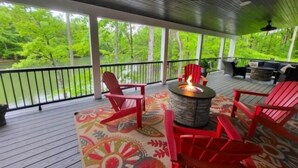 A covered outdoor deck with red Adirondack chairs, a central fire pit, and a view of a green landscape. - 17 ft Swim Spa, hot tub, pool table, kayaks, 3 kitchens, wifi (Moneta)