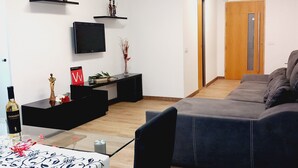 Un salon moderne avec un canapé gris, un téléviseur à écran plat fixé au mur et une petite table sur laquelle se trouvent une bouteille et quelques objets décoratifs.