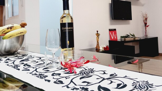 Une table à manger avec une bouteille de vin Canarius, un panier de bananes et un verre. On peut voir des objets décoratifs sur le mur et une petite statue sur une étagère.