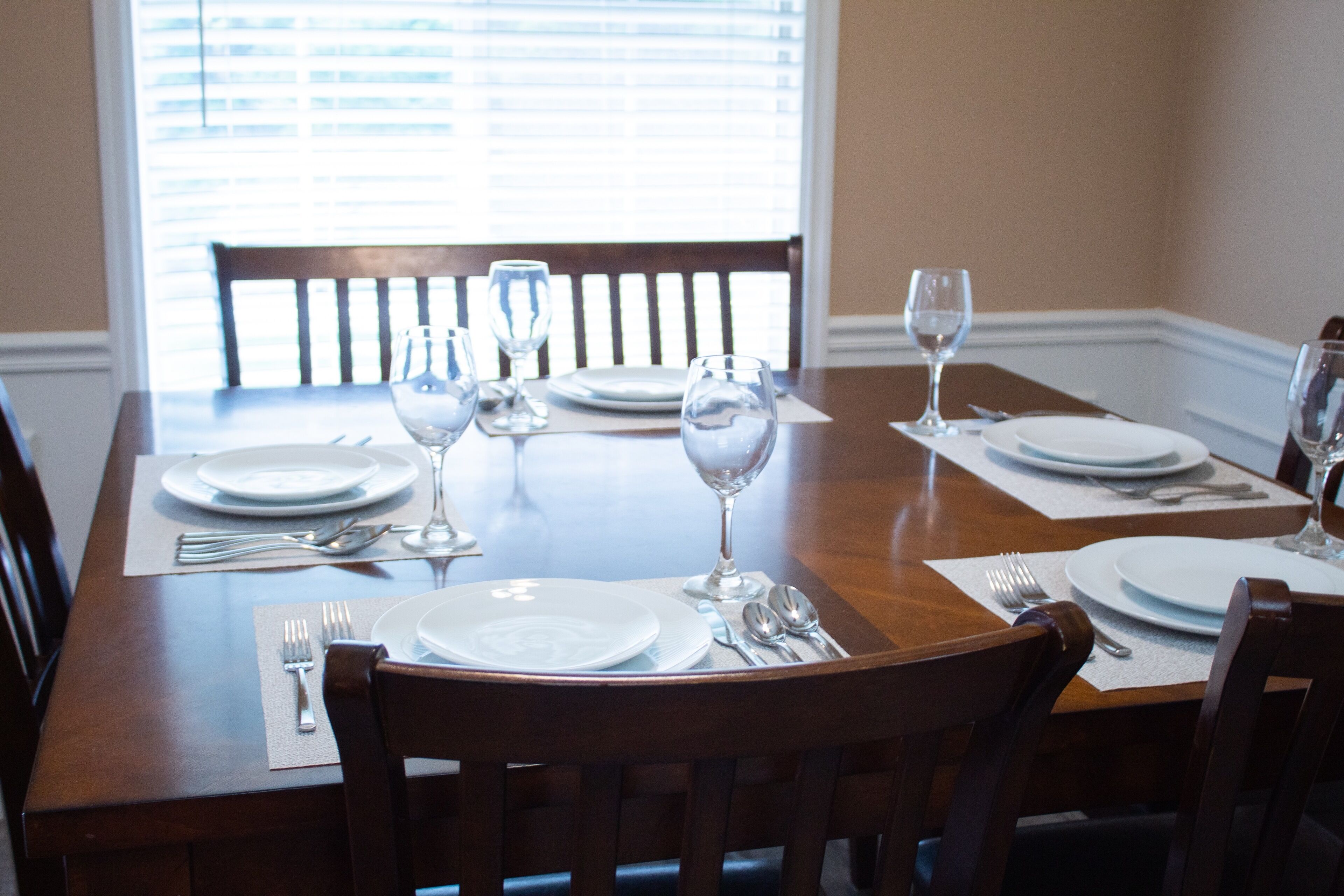A dining table set with plates, cutlery, and wine glasses, with a window in the background.
