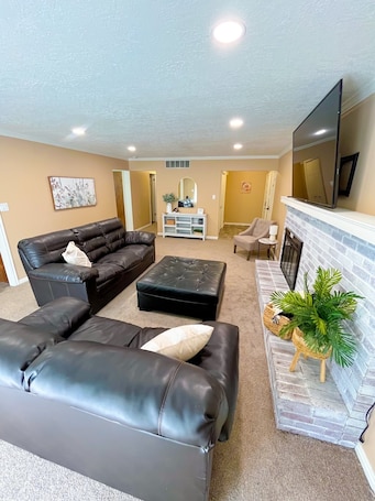 A living room with a black leather sofa, a matching ottoman, a fireplace, and a flat-screen TV mounted above it.