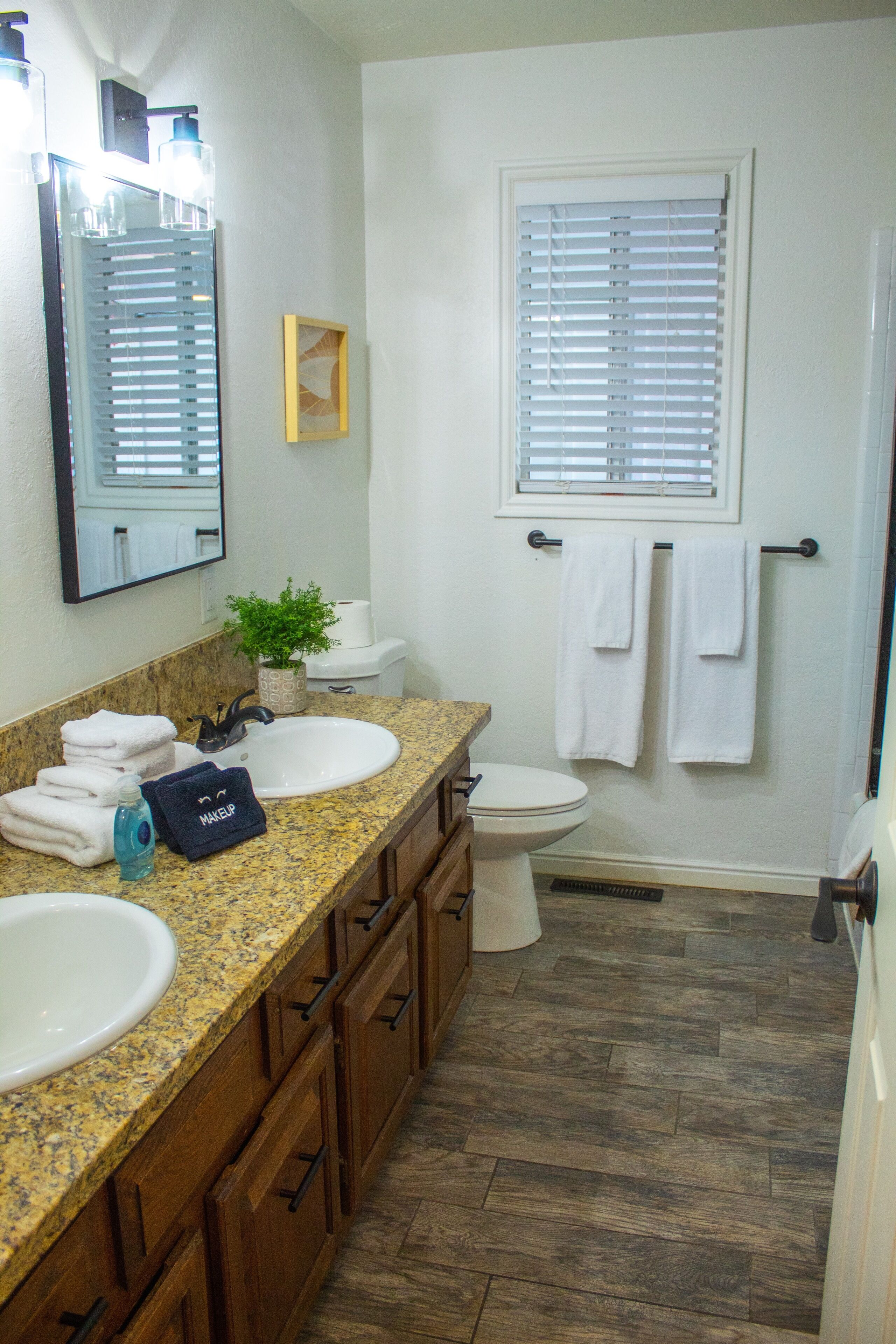 Une salle de bain avec deux lavabos, des toilettes et une fenêtre avec des stores.