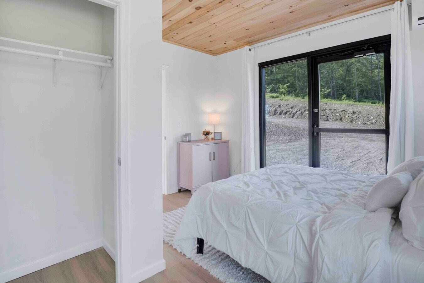 Een slaapkamer met een bed, nachtkastje, kast en een groot raam met uitzicht op een bos.