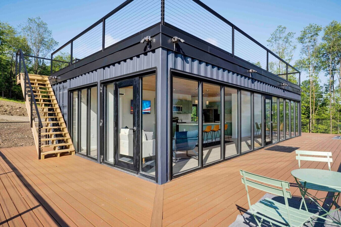 Een modern glazen huis met een houten terras, een trap en buitenmeubilair.