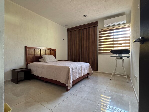 A bedroom with a wooden bed, a nightstand, a window with blinds, and an air conditioning unit.