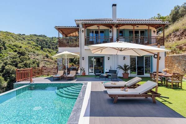 A two-story house with a swimming pool, outdoor furniture, and a view of the surrounding hills.