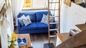 A cozy cabin interior with a loft bed, a blue sofa with patterned cushions, a wooden dining table, and a kitchen area.