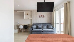 Et moderne soveværelse med en stor seng, en grå sofa, et skrivebord med stol og et fjernsyn ophængt på væggen.