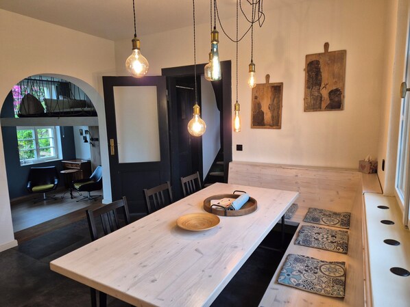 A modern dining area with a long wooden table, hanging light fixtures, and a view of a kitchen and dining space.