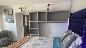 Een slaapkamer met een bed, een stoel, een kledingkast en een plank met decoratieve objecten.