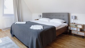 Una cama bien hecha con una colcha gris y almohadas blancas, una mesita de noche con una lámpara y una ventana con cortinas.