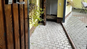 Una puerta de madera con los números 1 y 2, un sendero de ladrillo que lleva a una puerta y un pequeño espacio al aire libre con una silla y plantas en macetas.