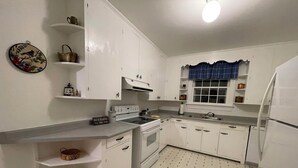 Una cocina con armarios blancos, estufa, fregadero y refrigerador. Hay repisas con cestas y un plato decorativo en la pared.