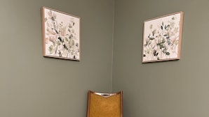 Una habitación de esquina con un sillón color mostaza, una mesita redonda con una planta y dos adornos florales en la pared.
