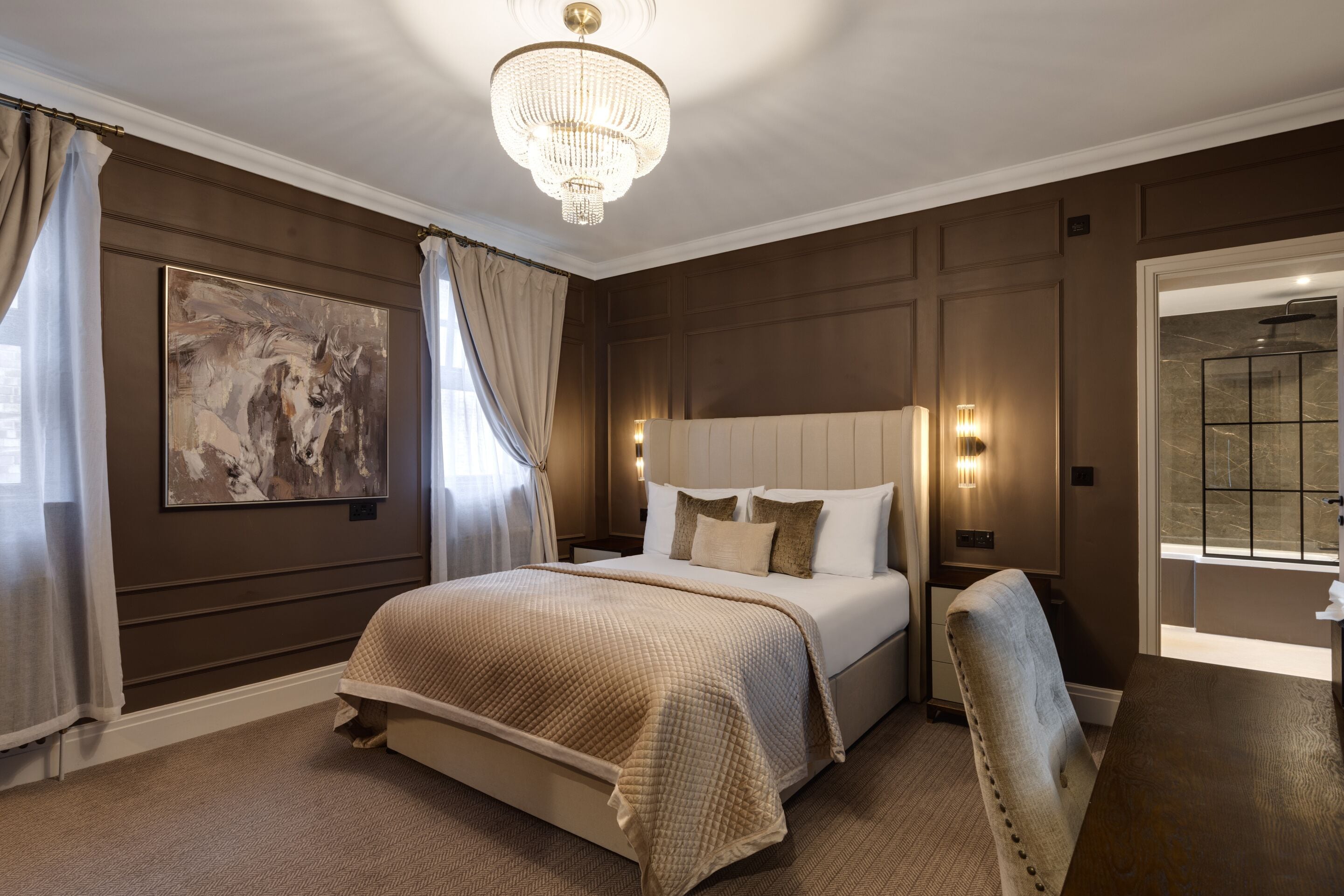 A bedroom with a large bed, a chandelier, a painting, and a bathroom visible through an open door.