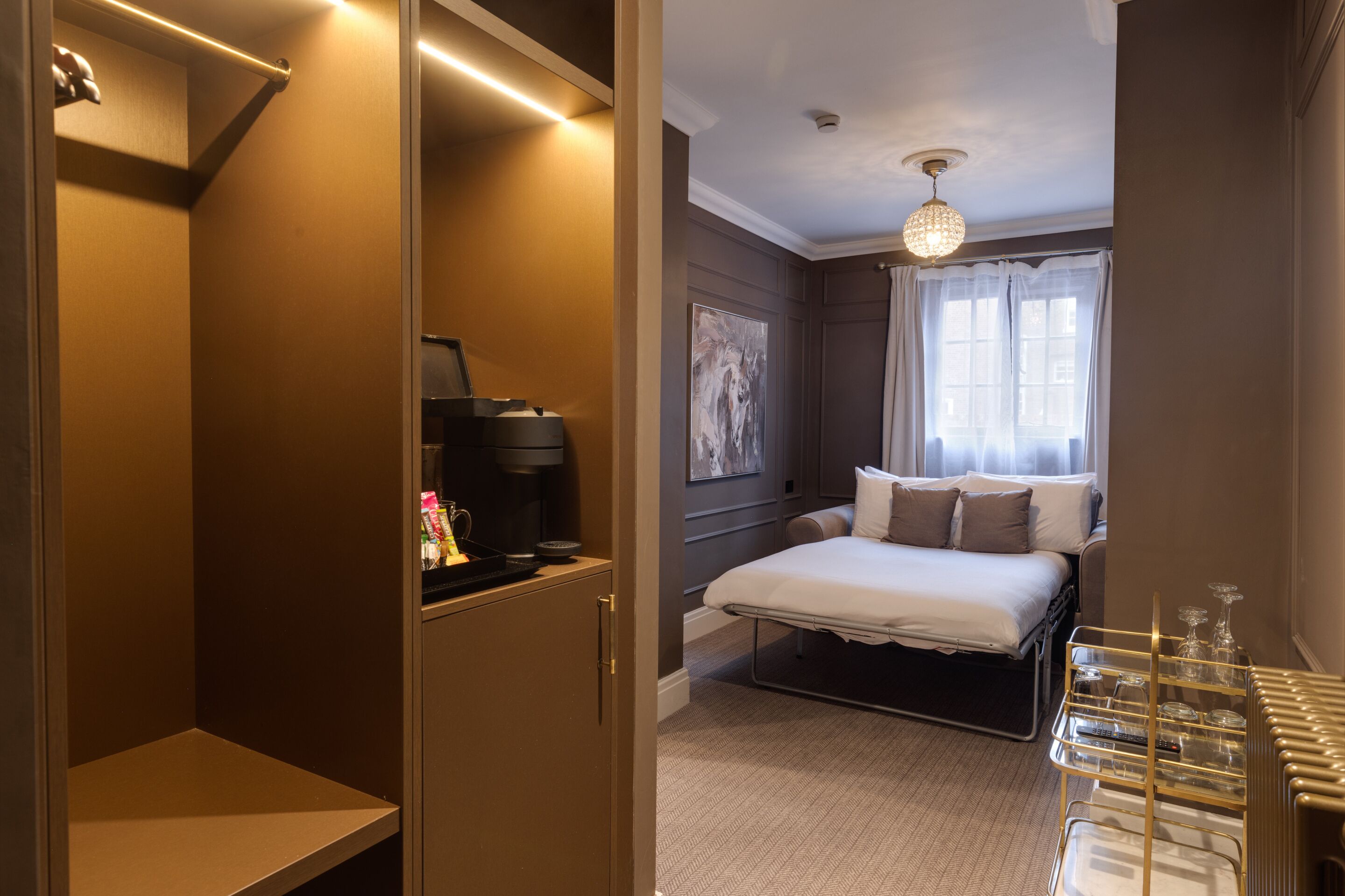 A hotel room with a bed, a bedside table, a coffee machine, a window with curtains, and a chandelier.