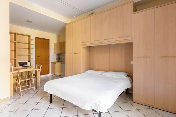A bedroom with a bed, wooden wardrobe, and a small kitchenette area with a microwave and chair.
