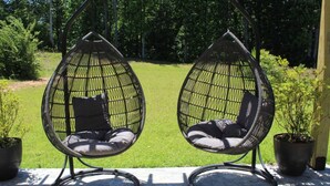Dos sillones colgantes con cojines en un patio con césped y árboles al fondo.