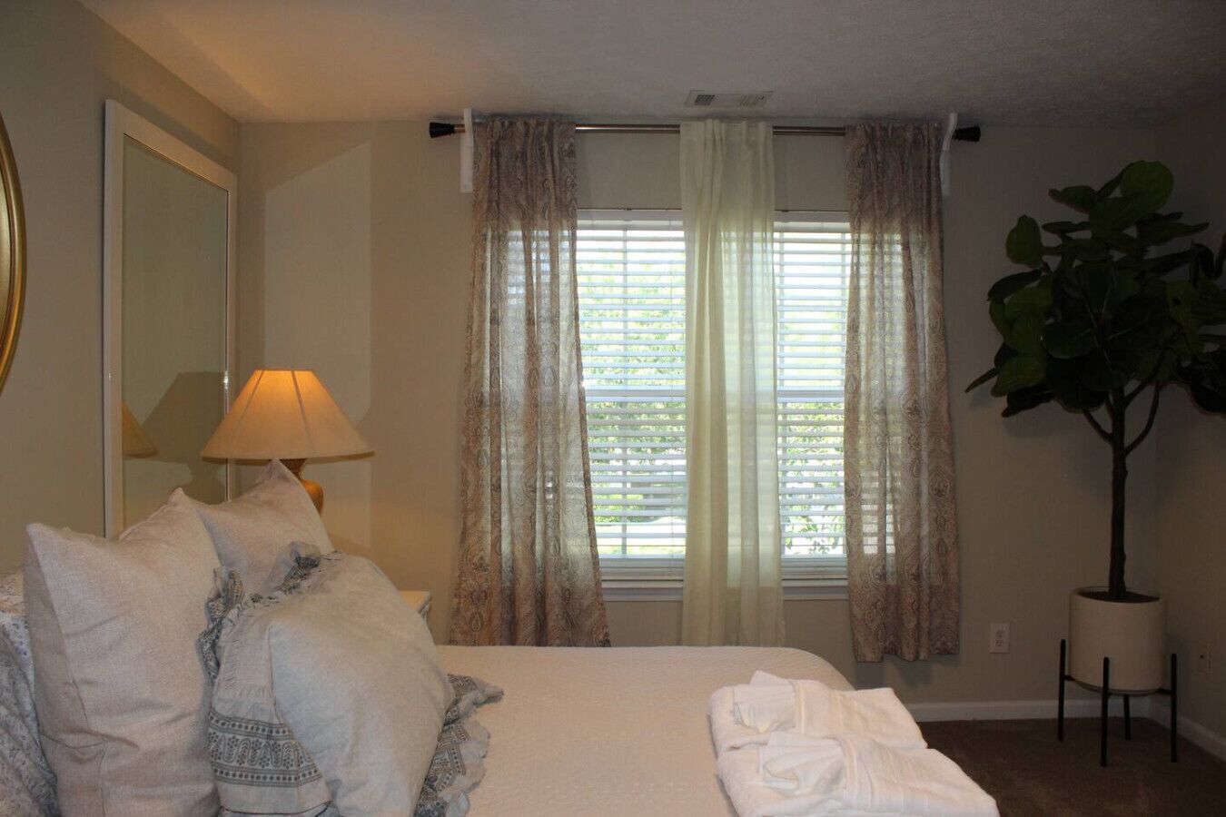 A hotel room with a bed, a lamp, a window with curtains, and a potted plant.