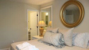 Habitación de hotel con una cama bien hecha, una lámpara de noche, un espejo grande y una puerta que da a un baño.
