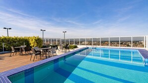 Ein Pool auf dem Dach mit freiem Blick auf die Stadt, umgeben von Sitzgelegenheiten im Freien und einer Glaskonstruktion.