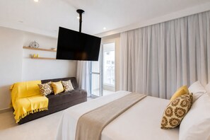 A modern hotel room with a large bed, a sofa, a flat-screen TV mounted on the wall, and a large window with curtains.