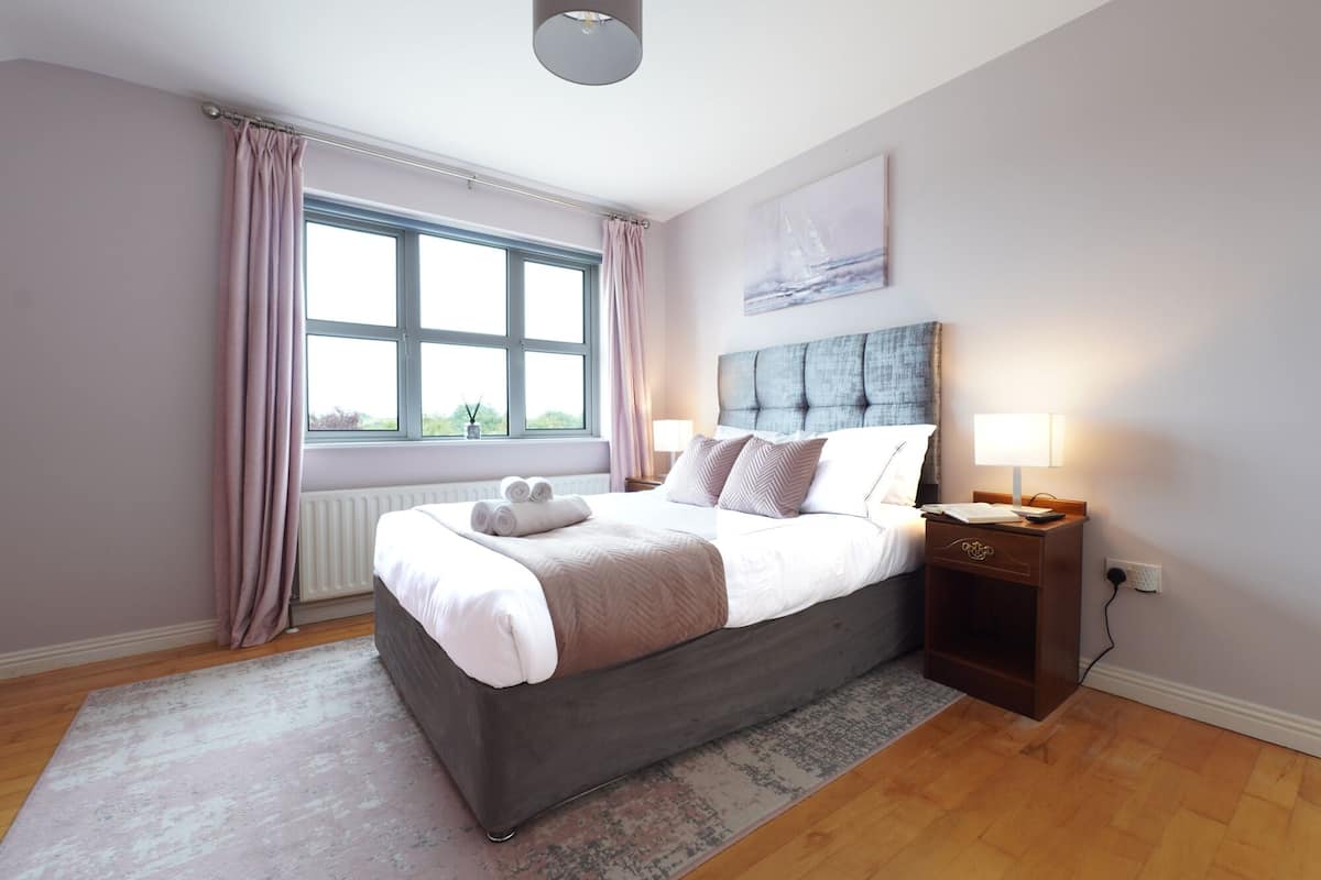 A bedroom with a large bed, a nightstand, a window with curtains, and a wall-mounted light fixture.