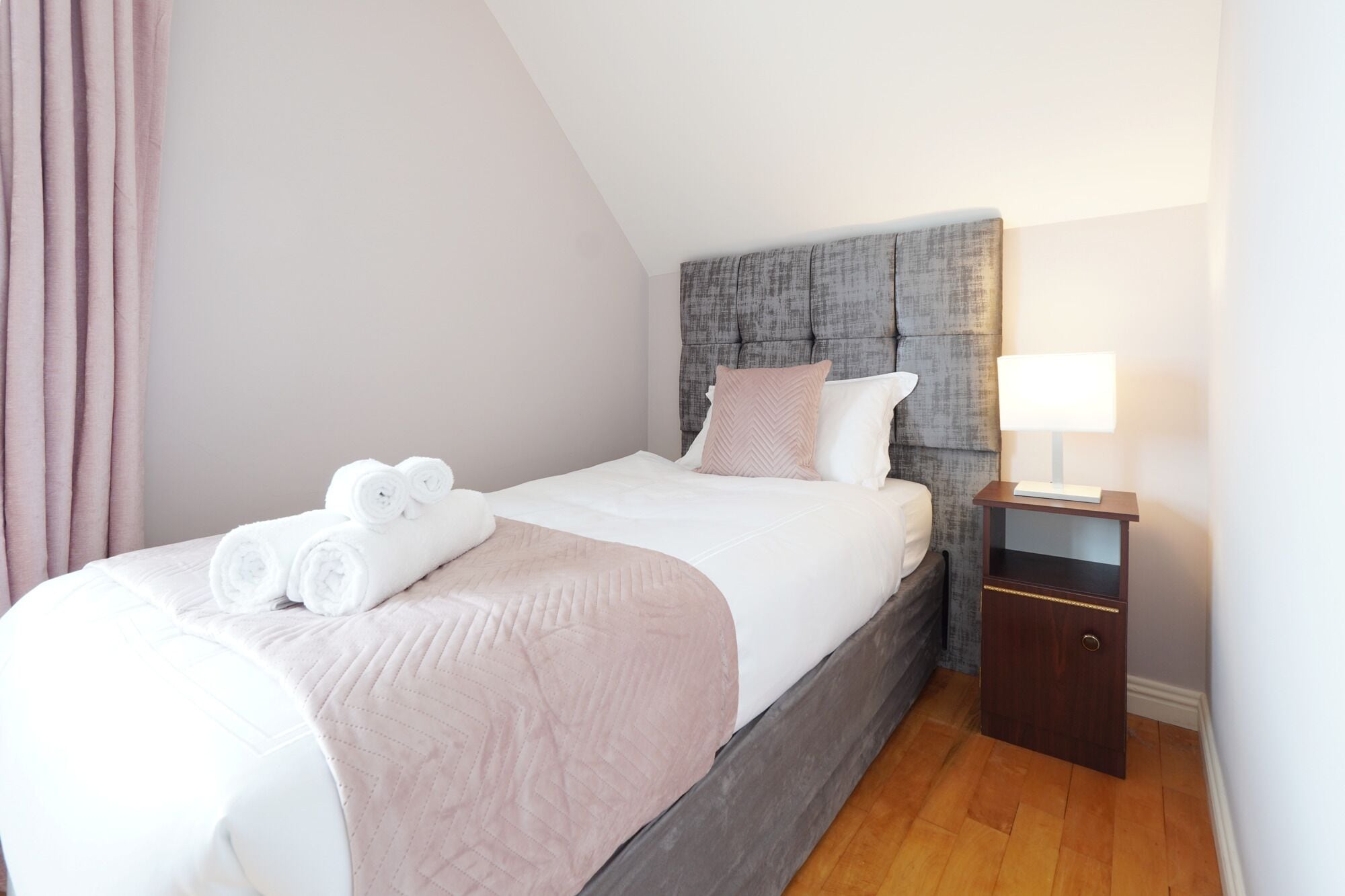 A neatly made bed with white linens and rolled towels, a grey upholstered headboard, a bedside table with a lamp, and a wooden floor.