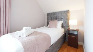 A neatly made bed with white linens and rolled towels, a grey upholstered headboard, a bedside table with a lamp, and a wooden floor.