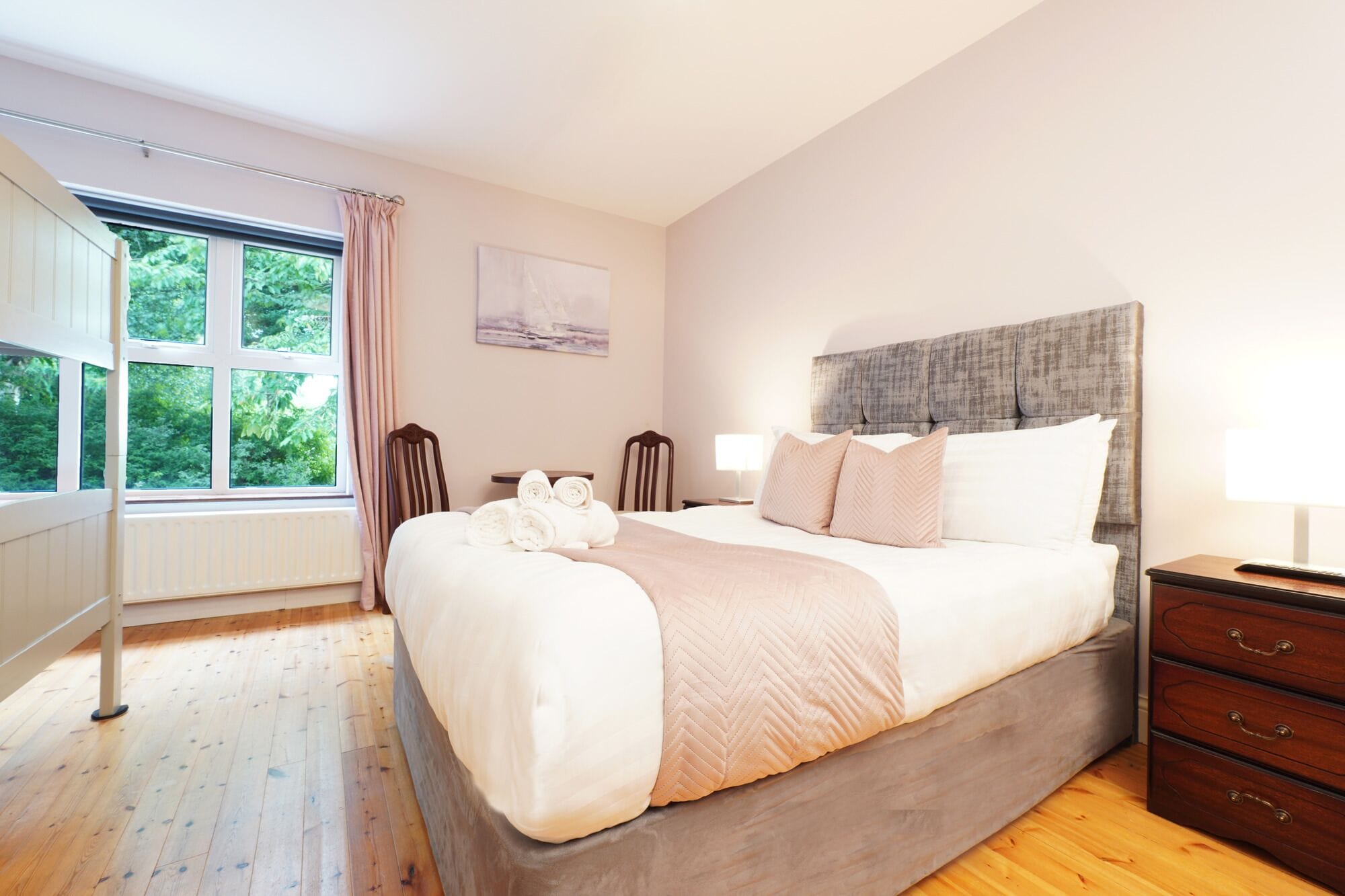 Un dormitorio con una cama grande, suelo de madera, una ventana con cortinas, cabecero y mesita de noche con lámpara.