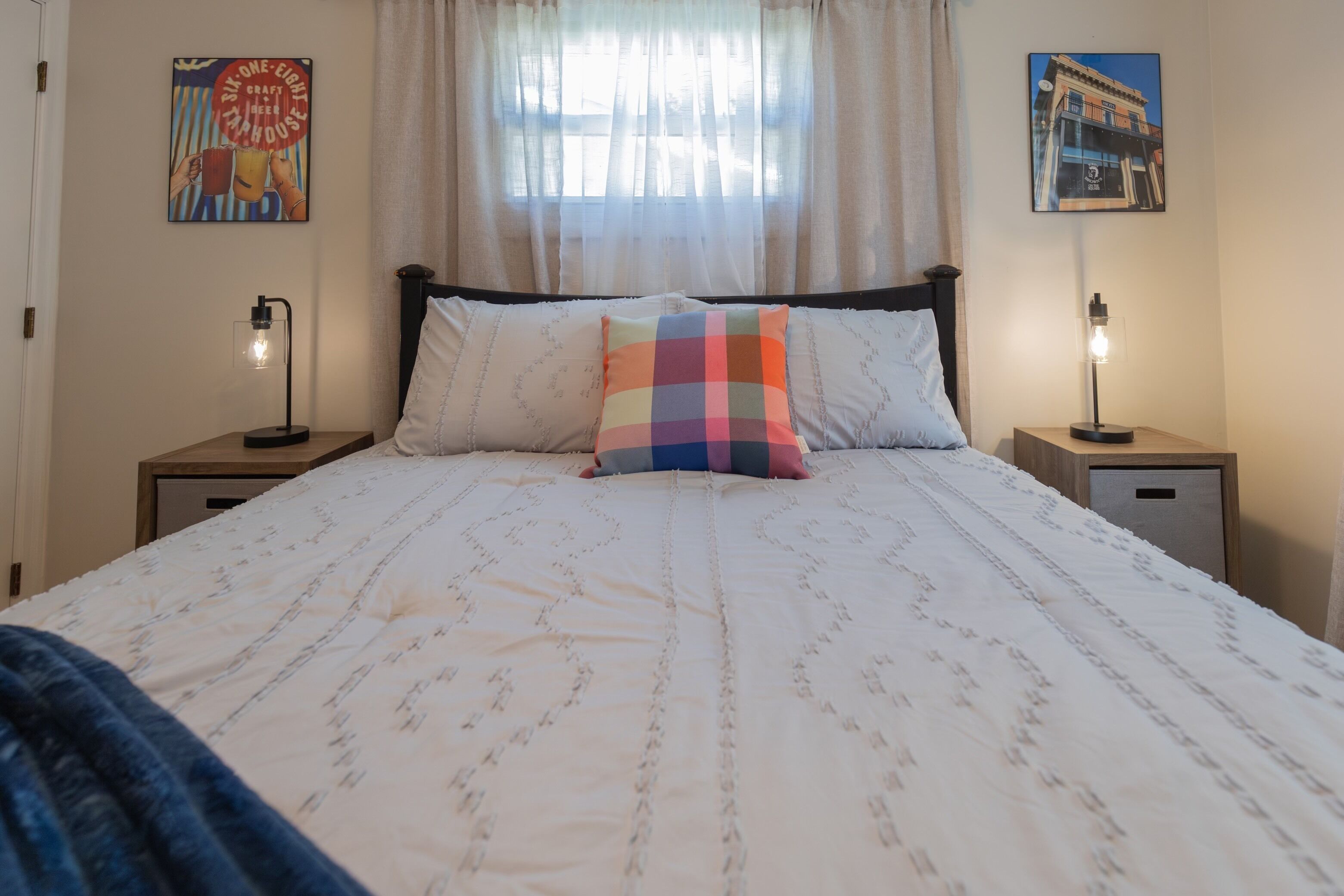 A neatly made bed with a patterned bedspread and a colorful pillow, flanked by two bedside tables with lamps, in a room with a window and two wall-mounted pictures.