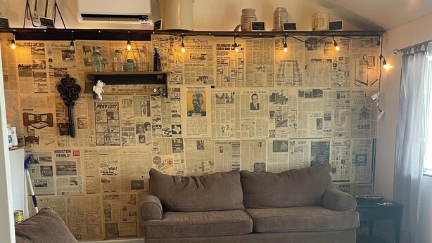 A living room with a sectional sofa, a small table, and a wall covered with newspaper clippings. - Nana’s Bunkhouse is a small little efficiency located in Houston, MO.  (Houston)