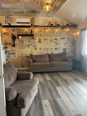 A living room with a sectional sofa, a small table, and a wall covered with newspaper clippings. - Nana’s Bunkhouse is a small little efficiency located in Houston, MO.  (Houston)