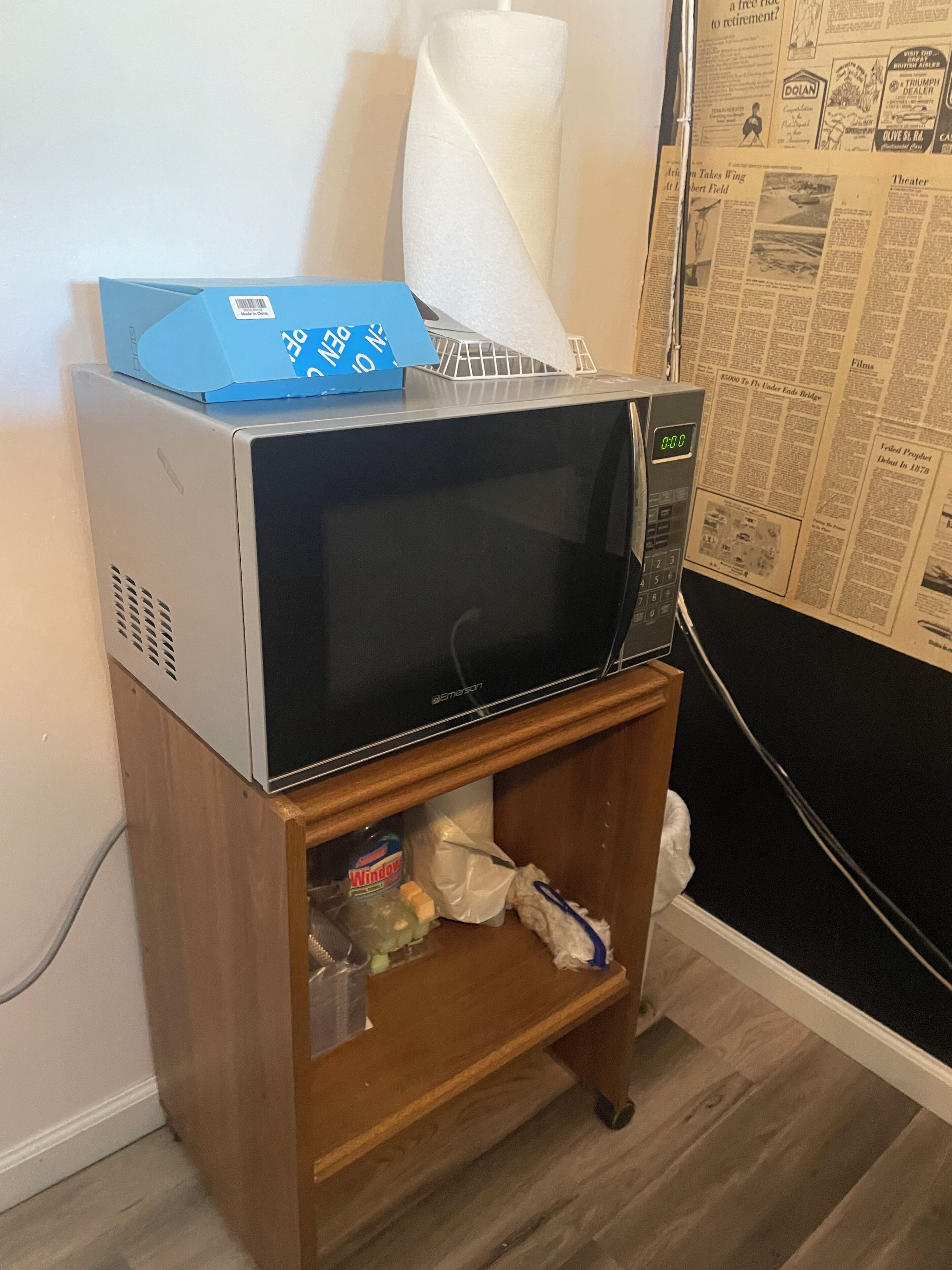 A microwave on a wooden shelf with a newspaper, a bottle of window cleaner, and a tissue box.