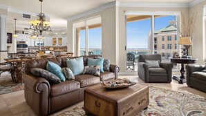 A spacious living room with a brown leather sofa, a wooden coffee table, and a patterned rug. A dining area with a wooden table and chairs is visible in the background. - Sweeping Gulf and Bay views on Clearwater Beach, 15th floor Penthouse 3 BR/3BA (Clearwater)