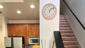 Eine moderne Küche mit Holzausbau, einem weißen Kühlschrank, einer Mikrowelle und einem Essbereich mit weißen Stühlen. An der Wand hängt eine Uhr, und ein Bambushalter ist sichtbar.