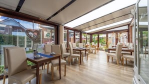 A spacious patio area with multiple tables and chairs, large windows offering views of the surrounding garden and buildings. - Pension Nordlicht (Esens)