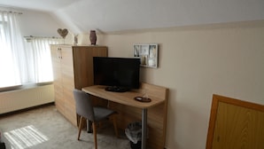 A room with a wooden desk, a television, a chair, a radiator, a window with curtains, and a wall with framed pictures. - Pension Nordlicht (Esens)