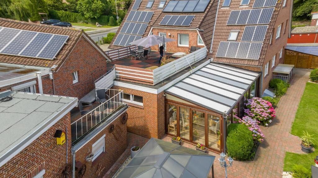 A residential building with multiple solar panels on the roof, a covered patio area, and a garden with flowers.