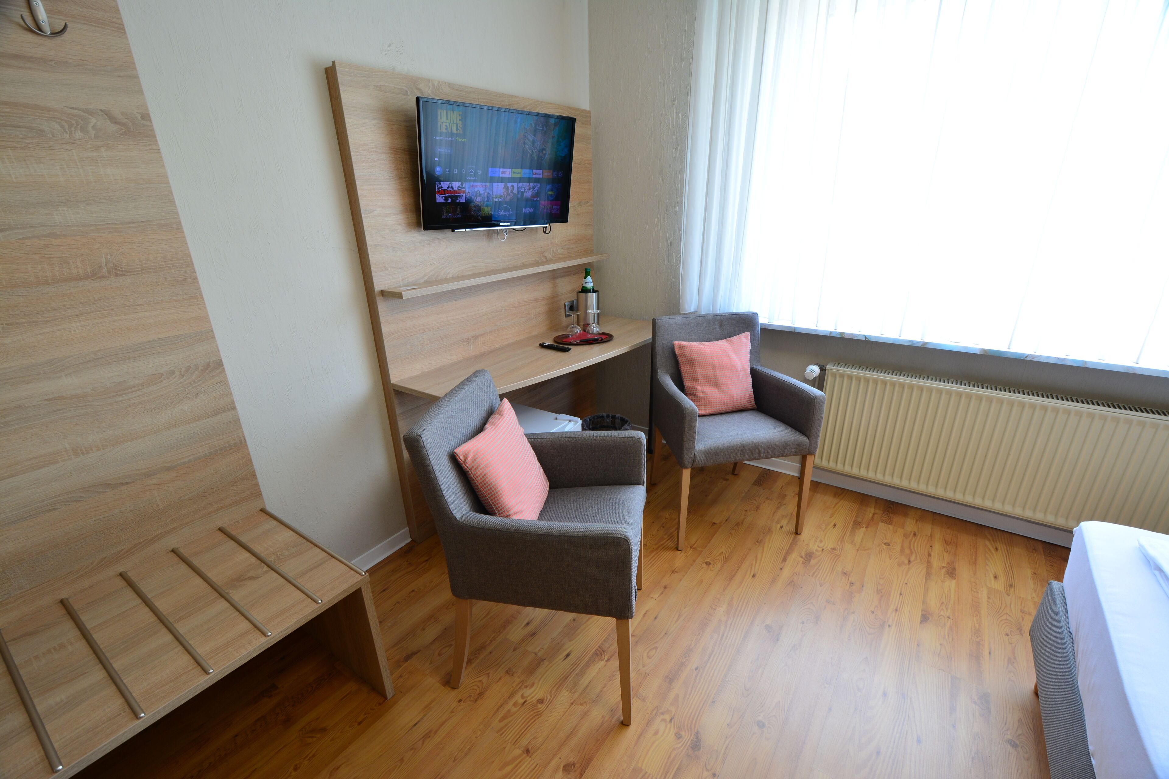 A modern hotel room with a wooden floor, a TV mounted on a wall, two grey armchairs with pink cushions, and a bed with a white duvet.